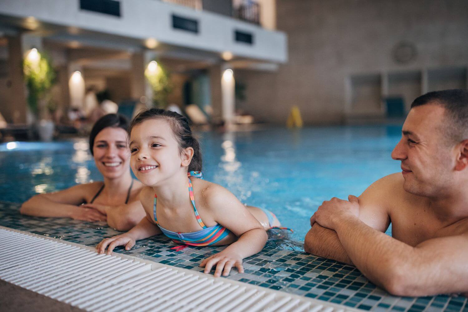 A young girl in a colorful swimsuit leans over the edge of a swimming pool, while two adults relax nearby.