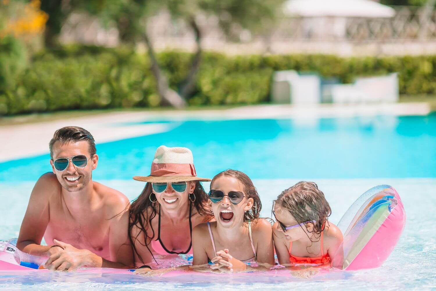 A family of four, wearing swimsuits, smiles together in a bright blue pool on a sunny day, showcasing a joyful summer vibe.