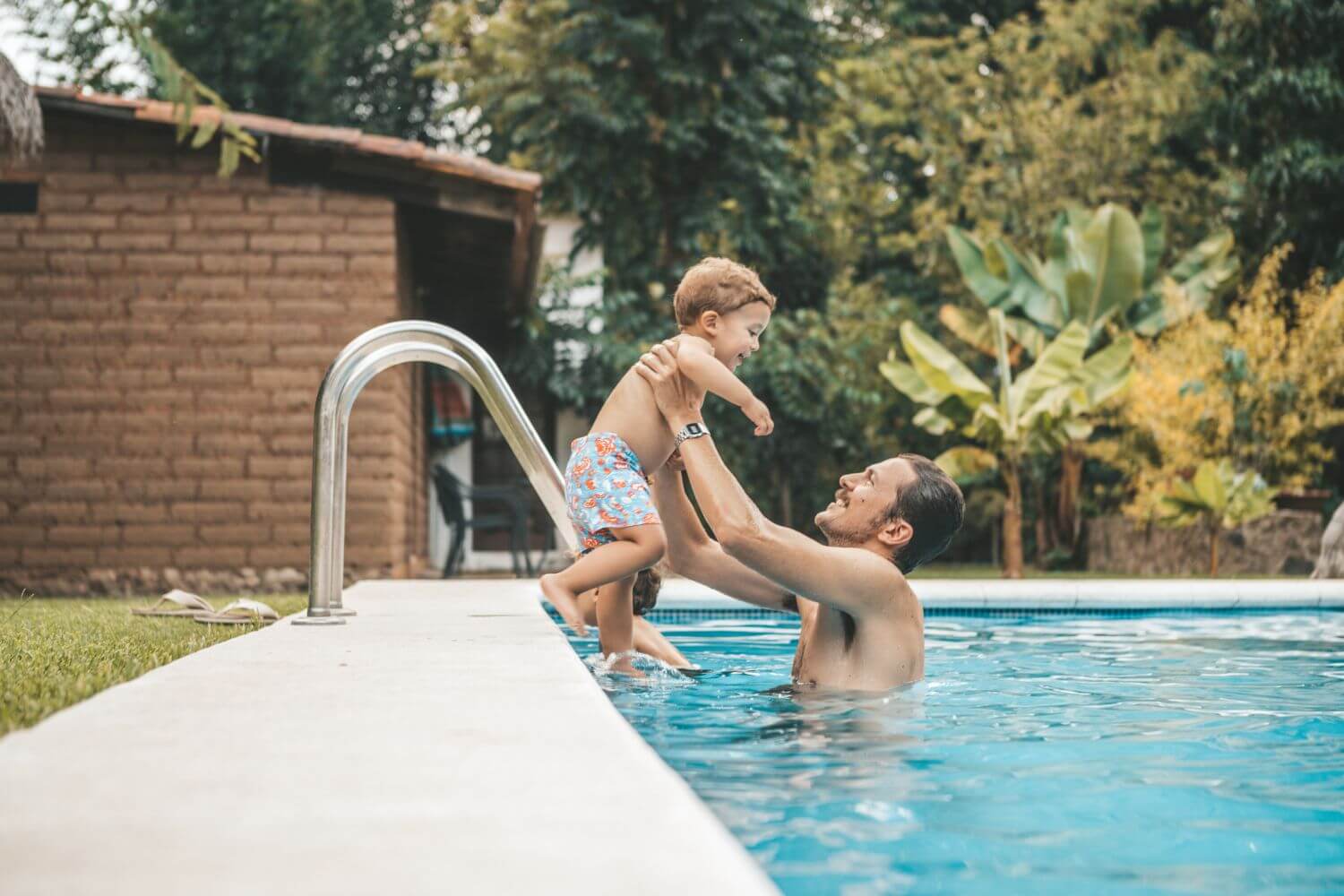 SiteWorx-Service-Areas-2 A man lifts a young boy out of a swimming pool, surrounded by greenery and a rustic brick building in the background.