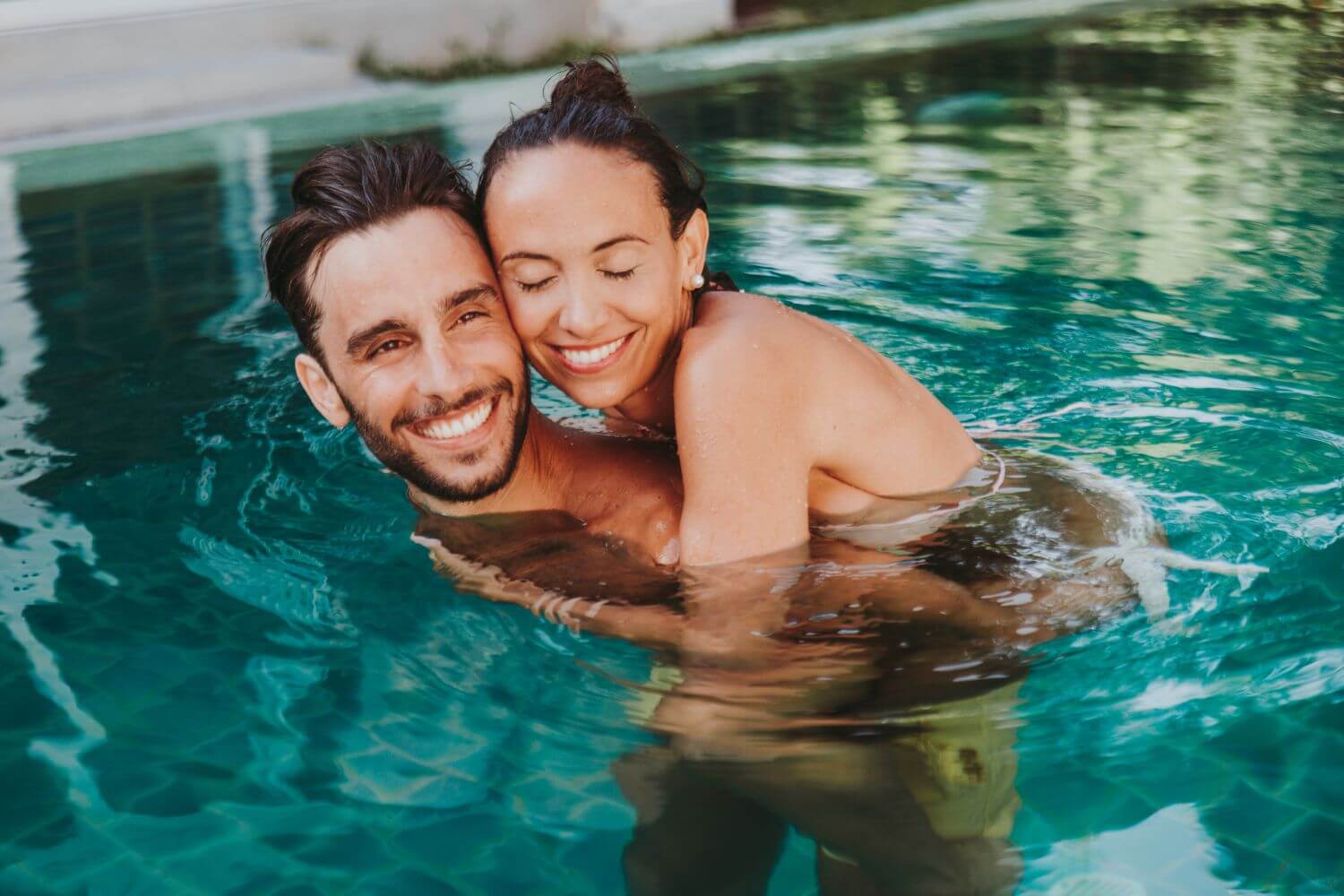 SiteWorx-Service-Areas-5 A couple enjoy a playful moment in a clear blue swimming pool, surrounded by lush greenery. Sunlight reflects on the water's surface.