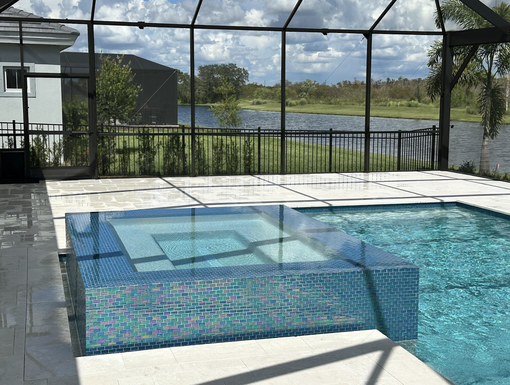 user-inserted A serene swimming pool with a raised, tiled spa overlooks a lush landscape and lake. The area is enclosed by a screened structure under a partly cloudy sky.