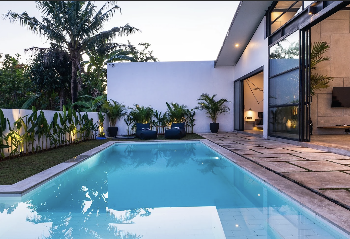 user-inserted Modern outdoor area with a serene pool surrounded by lush tropical plants. Warm lighting and open living space create a calm, inviting atmosphere.